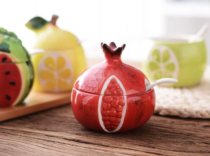 Ceramic sugar bowls shaped like fruits, including pomegranate, watermelon, lemon, and lime, complete with spoons, arranged on a wooden table