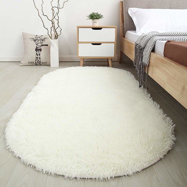A white, oval-shaped shaggy rug placed beside a bed.