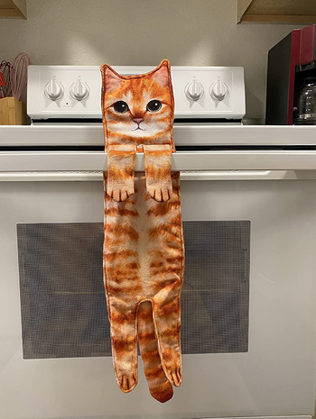 A towel shaped like a playful orange cat, with its paws draped over a stove handle, ready for a bit of fun in the kitchen.