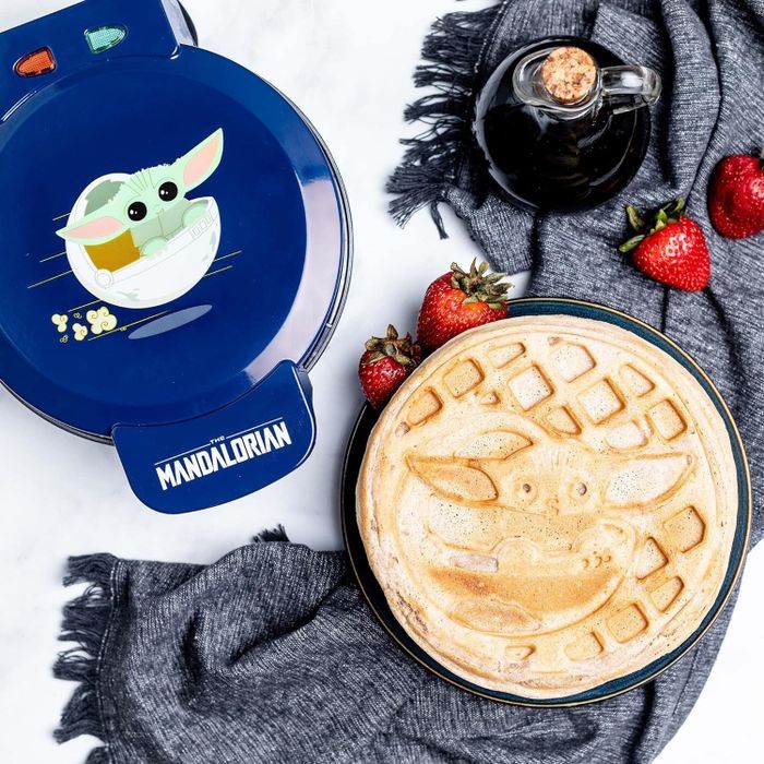Mandalorian-themed waffle maker with a Baby Yoda design next to a Grogu waffle, a bottle of syrup, and strawberries resting on a gray napkin