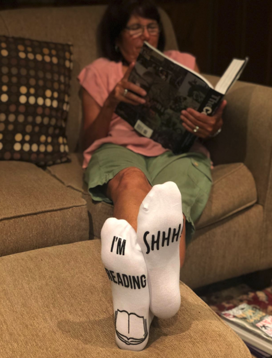 A reviewer’s image featuring a reader lounging with a book, feet up, proudly sporting the 'Shhh I'm Reading' socks.