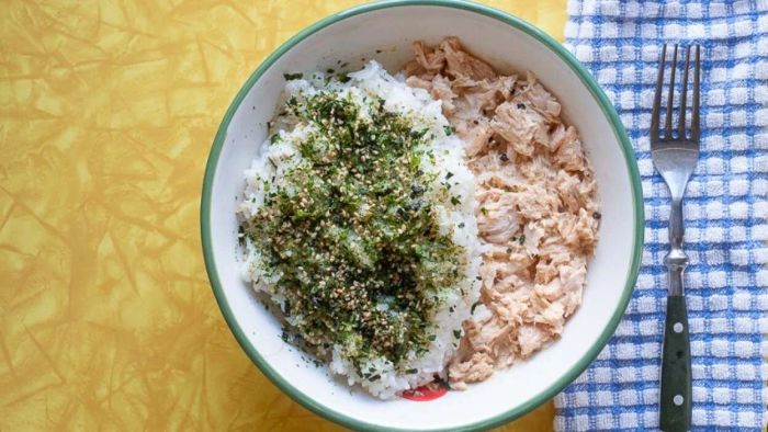 A top-down view of a rice bowl with tuna, topped with furikake, resting on a yellow table with an abstract design. Beside it, a blue-and-white checked napkin and a fork complete the scene.