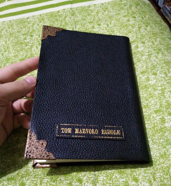 A close-up of a person holding a sleek black leather-bound journal adorned with gold corner details, embossed with 'Tom Marvolo Riddle' on the cover.