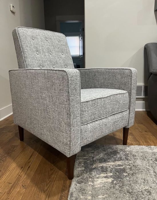 A gray upholstered armchair with button detailing on the backrest, complemented by wooden legs and a modern aesthetic, placed on a hardwood floor next to a textured rug.