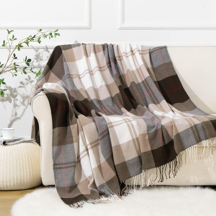 A plaid blanket casually draped over a beige couch, with a plant beside it and a pouf resting on the floor, creating a laid-back yet inviting space.