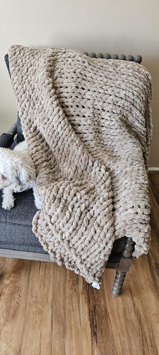 A small white dog sitting on a chair, partially covered by a textured blanket, looking cozy and content.