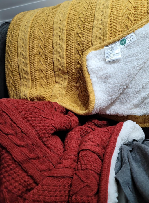 Two knitted blankets with plush, fluffy linings rest on a dark surface, highlighting their rich texture. One of the blankets has visible tags attached.