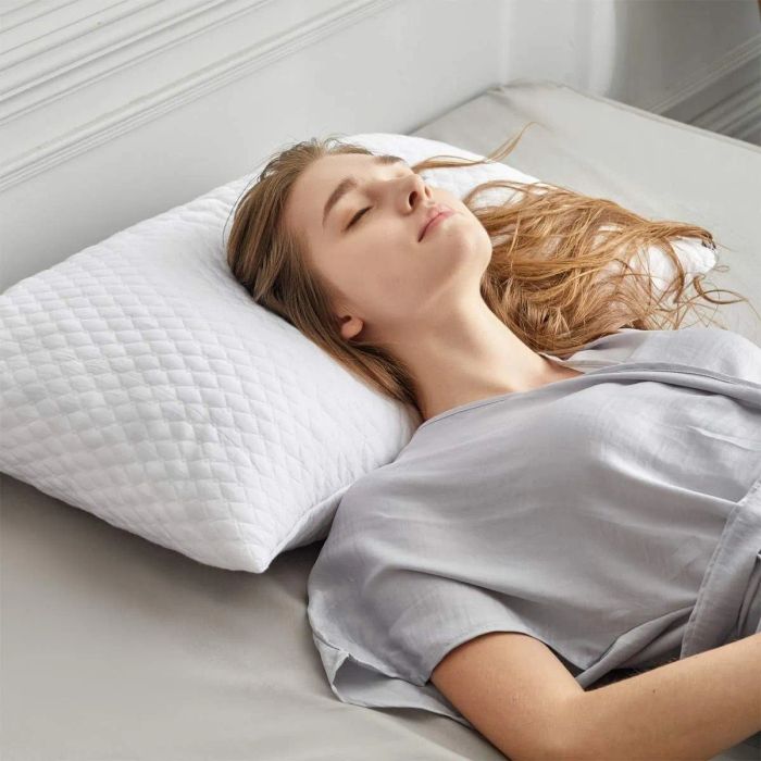 A woman peacefully resting on a bed with a white quilted pillow, wearing a light-colored shirt.