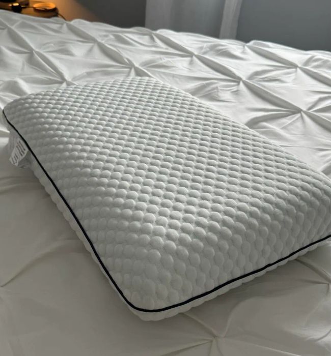 A textured pillow rests atop a neatly arranged bed with a white patterned comforter. A lamp and a window, revealing the outdoors, are visible in the background.