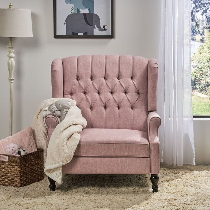 A wingback armchair with a tufted backrest, upholstered in fabric, situated in a cozy living room next to a floor lamp, a window, and a woven basket holding a blanket.