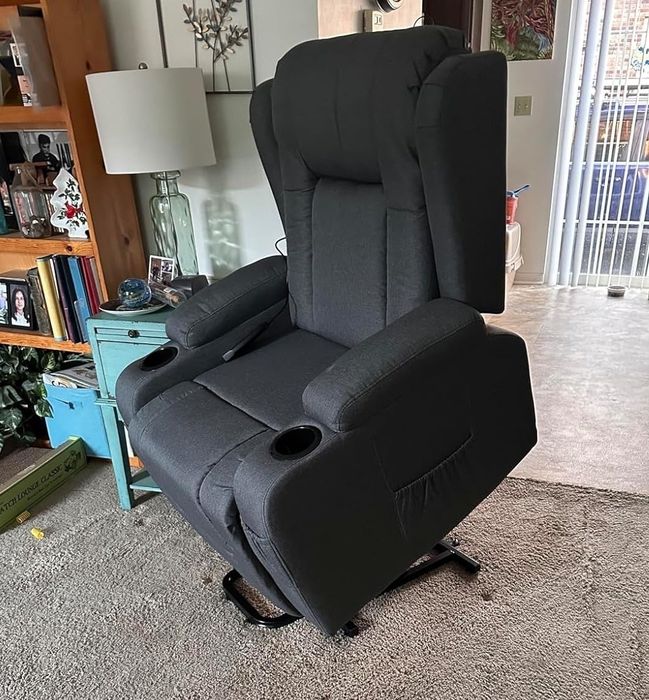A black power lift recliner chair is shown in a living room, with a lamp, teal nightstand, bookshelf, and a sliding door visible in the background.