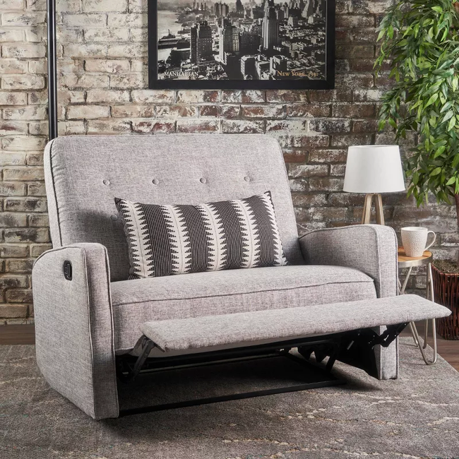 A gray loveseat recliner with a patterned cushion, placed against a brick wall decorated with a New York City poster. The setup includes a lamp, a plant, and a side table with a mug.