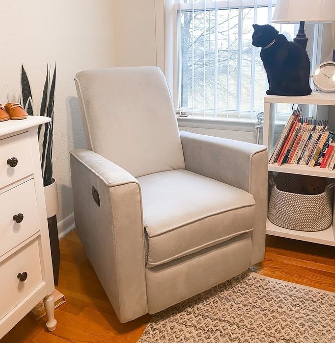 A light gray reclining chair in a cozy room, accompanied by a white wall sign. A black cat sits on a white table with books and a lamp, positioned by the window.
