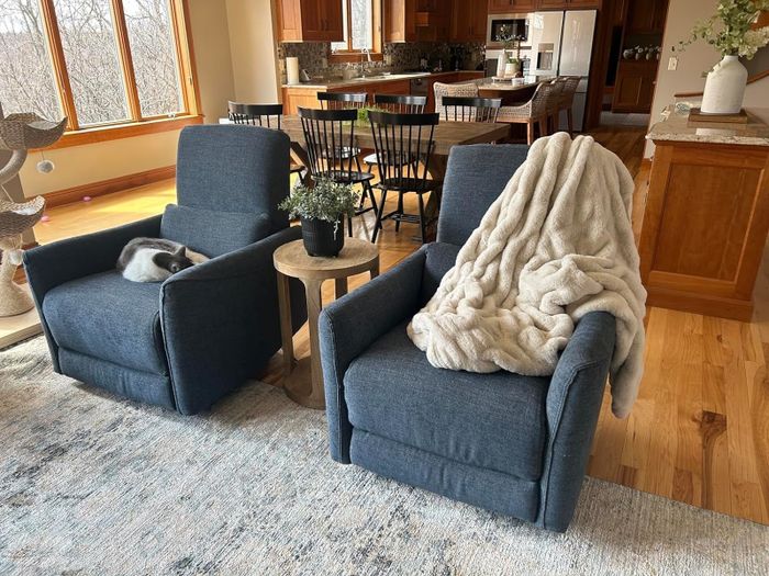 Two gray recliners in a warm and inviting living room. One is draped with a soft blanket, while a cat naps on the other. The space also features a dining area and a kitchen in the background.