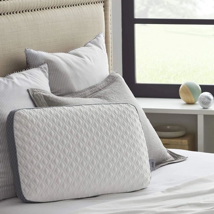 A quilted memory foam pillow placed on a neatly made bed with other decorative pillows, set in a cozy bedroom next to a window that frames a green outdoor view.
