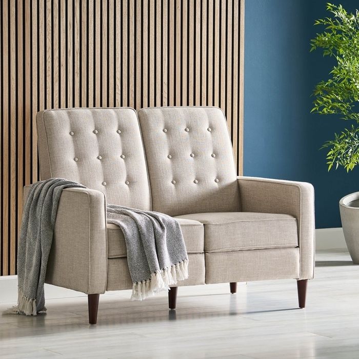 A modern beige loveseat featuring tufted backrests, dark wooden legs, and a gray fringed throw draped over one side. Positioned against a rustic wood-paneled wall.