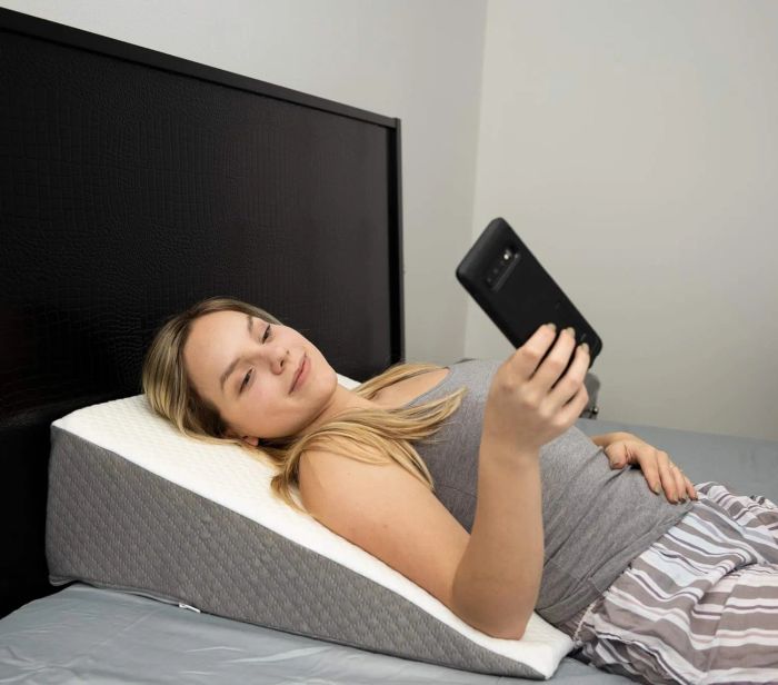 A person in a gray tank top and striped pajama pants is lying on a wedge pillow in bed, holding a smartphone.