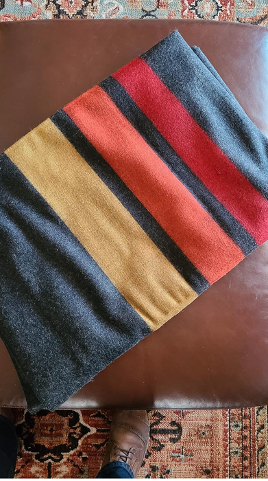 A folded wool blanket with bold stripes on a leather surface, accompanied by part of a person’s shoe near a patterned rug.