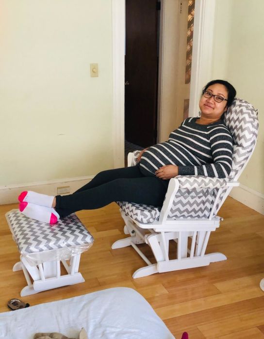 A pregnant woman in a striped shirt sits comfortably in a rocking chair with a matching footrest, relaxing with her feet up. She wears glasses and pink-tipped socks in a cozy room setting.