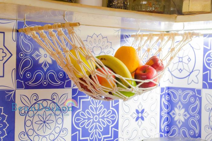 A macrame fruit hammock hanging from under a kitchen cabinet, filled with fresh fruit