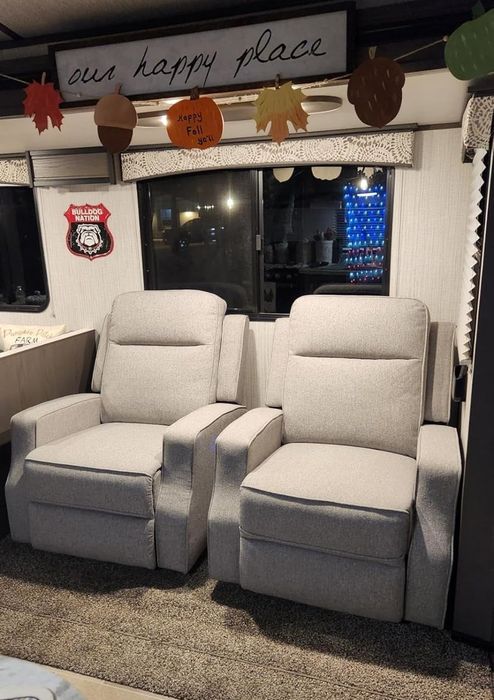 Two gray recliners side by side in a cozy living room, with a sign above that reads 'our happy place' and fall decorations hanging nearby. The room has carpeted flooring.