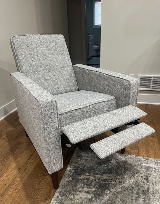 A modern gray fabric upholstered recliner with an extended footrest, positioned on a wooden floor next to a textured area rug.