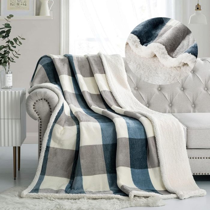 A cozy plaid fleece blanket casually draped over a sofa in a warm living room, complete with a plant and wall shelf in the background.