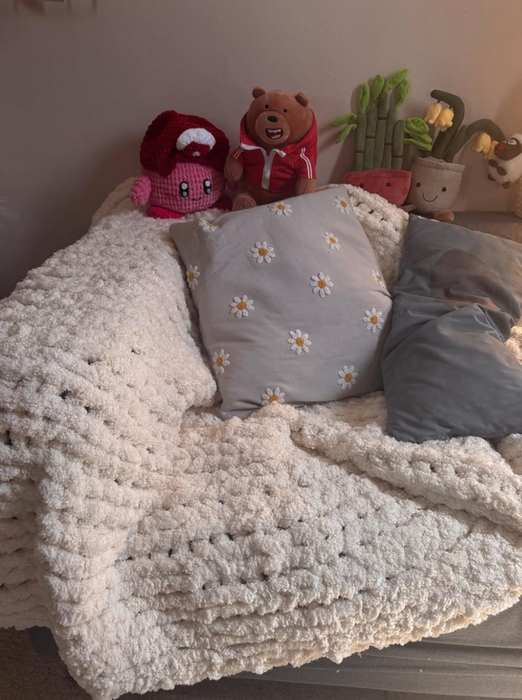 A cozy corner featuring stuffed toys, a crocheted mushroom, a bear in a hoodie, and pillows with floral patterns on a soft, textured blanket.