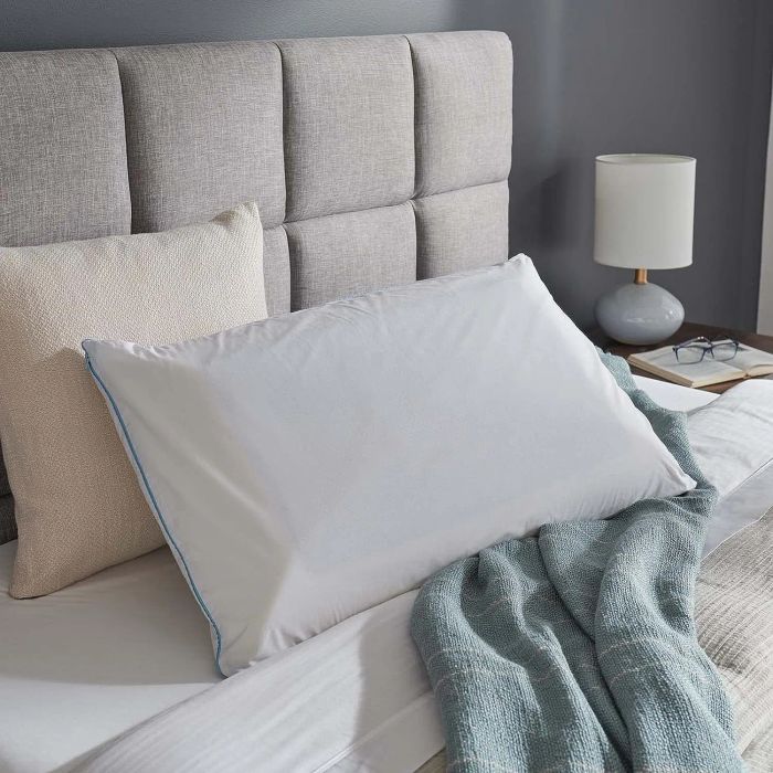 A cozy bedroom scene featuring a neatly made bed with a white pillow trimmed with blue piping, a beige pillow, a blue blanket, and a grey upholstered headboard. A small lamp and a pair of glasses resting on a book on a side table complete the setting.