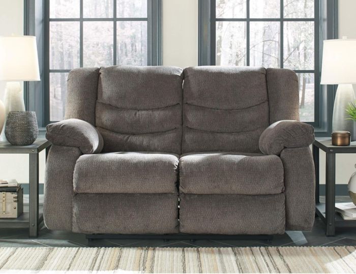A cozy gray recliner loveseat set in a comfortable living room, featuring large windows and side tables with lamps.