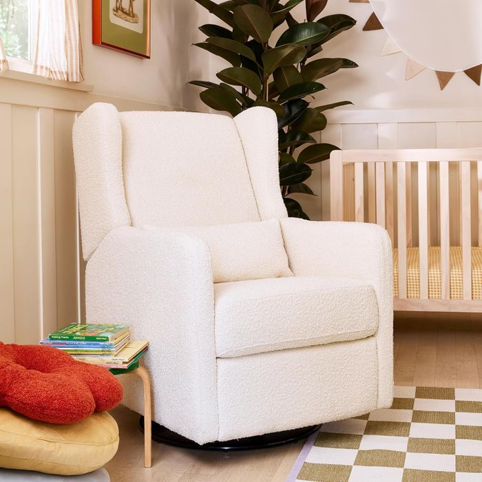 A modern nursery featuring a cozy white armchair, a wooden crib, a small table stacked with children's books, a cushion shaped like a paw, and a lush green houseplant.