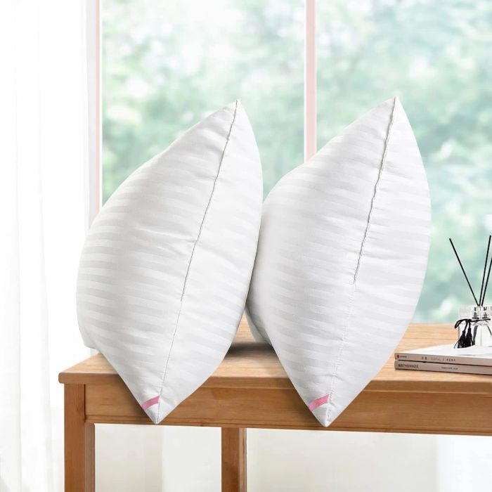 Two white pillows with delicate stripes rest against a window backdrop, positioned on a wooden table next to a reed diffuser.