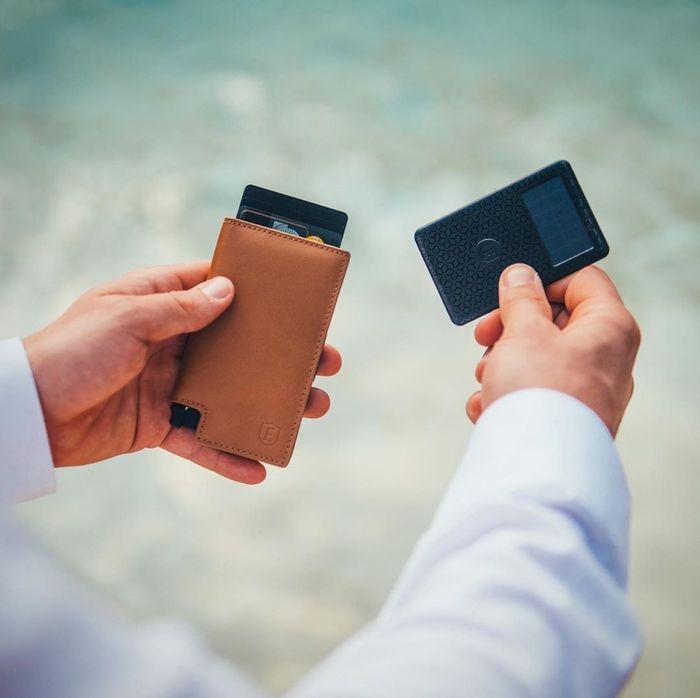 The rectangular light brown leather wallet in a model's hand, with cards peeking out of the top and the card-shaped tracker held in the other hand