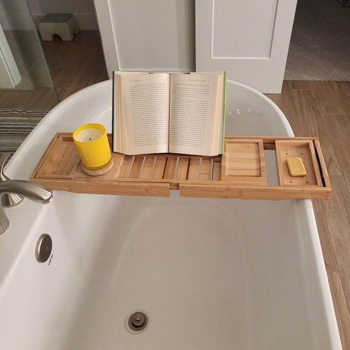 A bathtub caddy with a yellow candle, a book, and a bar of soap resting across a bathtub, evoking the ultimate bath-time relaxation experience.