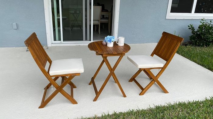 A cozy outdoor patio featuring a small wooden table and two matching chairs with white cushions. The table is adorned with blue flowers and two white mugs