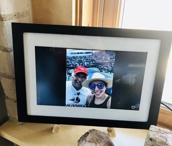 The frame with a white and black border around the digital screen, placed in the author's parent's home, displaying a picture of her and her father at a tennis match.