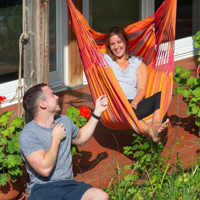 A man in a gray T-shirt and a woman in a hammock share a smile in a garden setting, surrounded by plants and a wooden house in the background