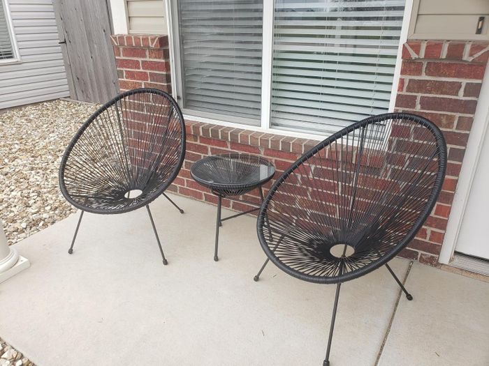 Two black circular wicker chairs and a small round table are set up on a patio in front of a window with blinds