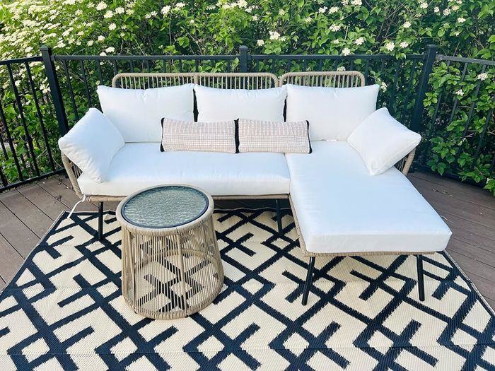 A patio setup featuring a white cushioned sectional sofa and a glass-top wicker table placed on a patterned outdoor rug, surrounded by lush greenery