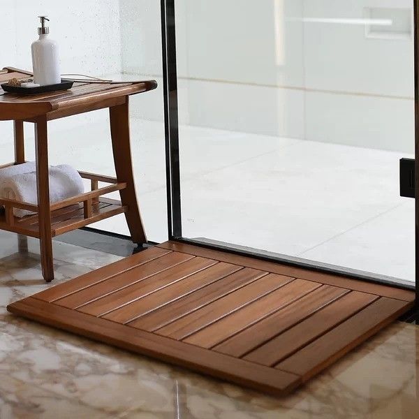 A bathroom scene featuring a wooden slatted bath mat, a small table with a towel and soap dispenser placed next to a glass shower door on a sleek marble floor.