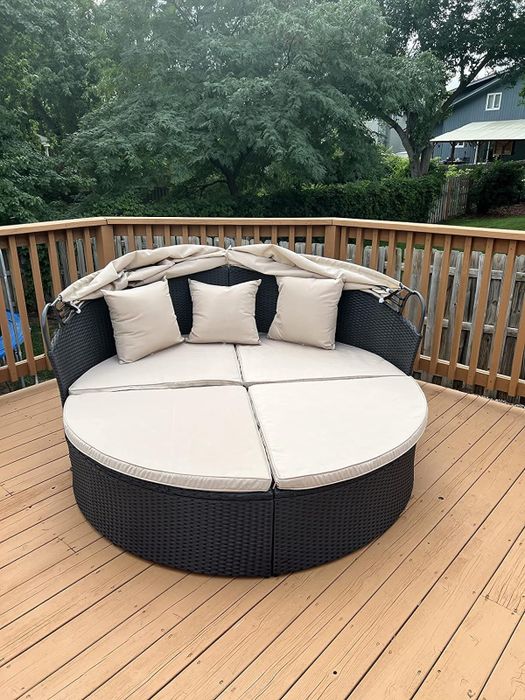 A round rattan daybed with beige cushions sits on a wooden deck outdoors. It features an adjustable canopy and four decorative pillows, with trees and houses visible in the background