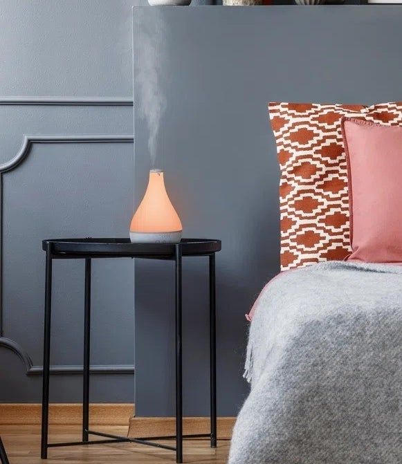 A contemporary bedroom with a neatly arranged bed layered with pillows, a sleek nightstand that holds a diffuser, and a decorative shelf above adorned with vases and artwork.