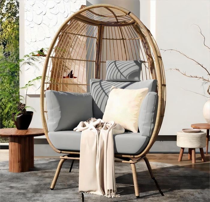An oval-shaped rattan hanging chair featuring gray cushions and a cream throw blanket, perfect for creating a snug reading corner