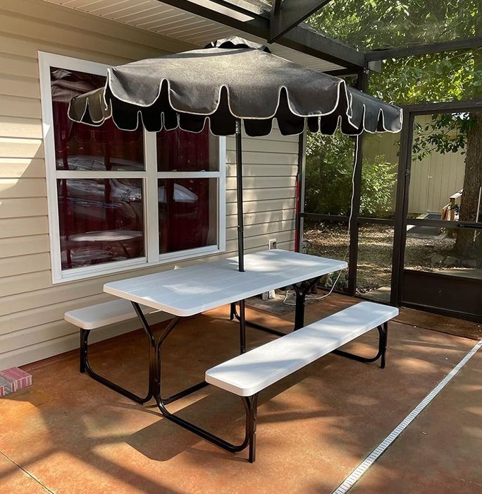 An outdoor patio setup with a white picnic table and attached benches under a black umbrella with white trim, positioned near a house window