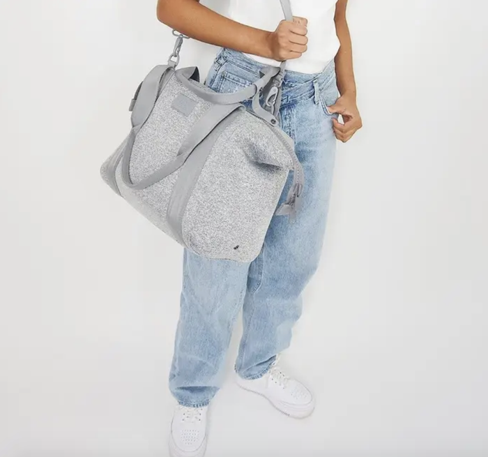 Model wearing the long strap of the grey square-shaped duffel with shorter handles