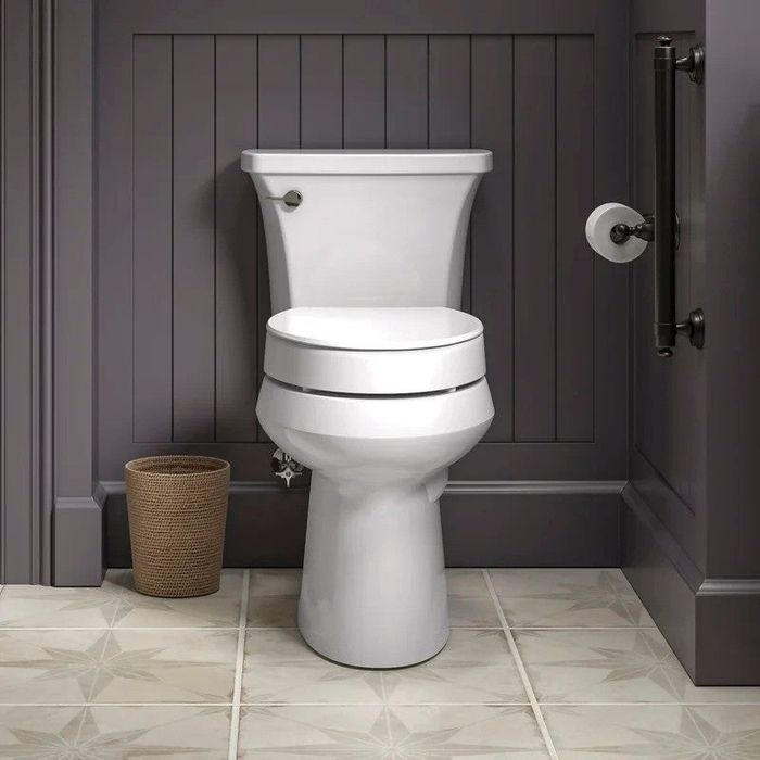 A white toilet positioned against a paneled wall, with a small wicker wastebasket on the left and a toilet paper holder on the right side.
