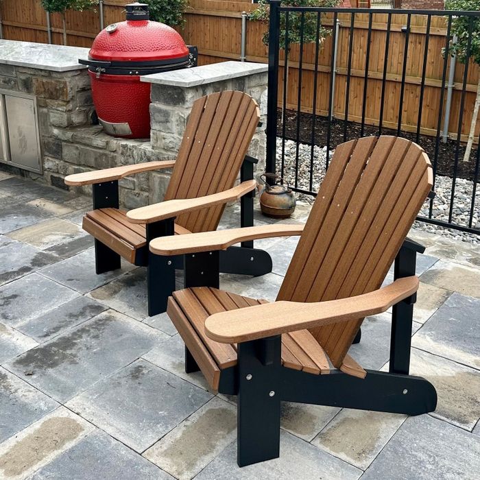 Two wooden Adirondack chairs with black frames are positioned on a stone patio beside a red kamado grill and a stone outdoor kitchen