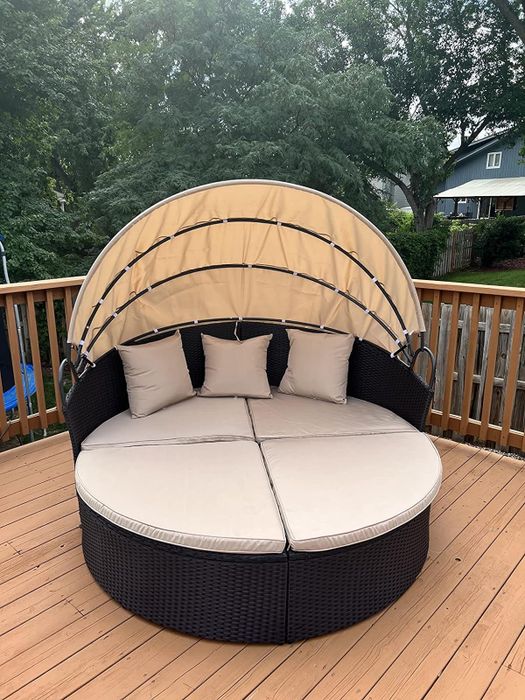 A round outdoor daybed with a retractable canopy sits on a wooden deck, adorned with beige cushions and pillows, set against a backdrop of trees and a house