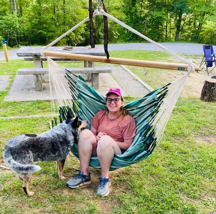 A woman casually dressed in shorts, a t-shirt, a red cap, and glasses sits on a hanging hammock outdoors, with a dog standing nearby