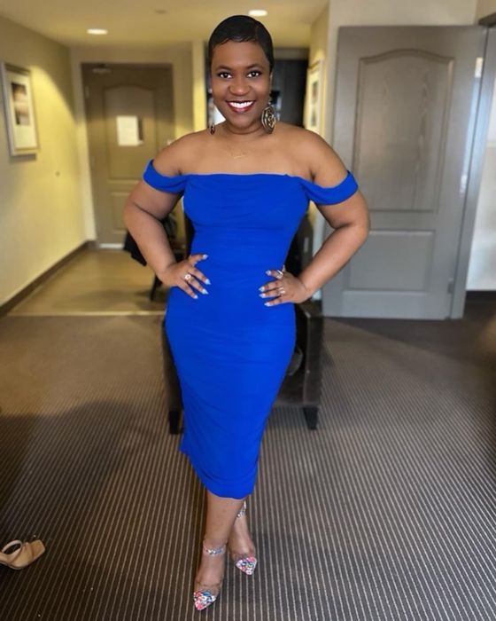 A reviewer confidently posing in a flattering off-shoulder blue dress with a form-fitting silhouette, paired with bold earrings and open-toe heels, smiling gracefully indoors.